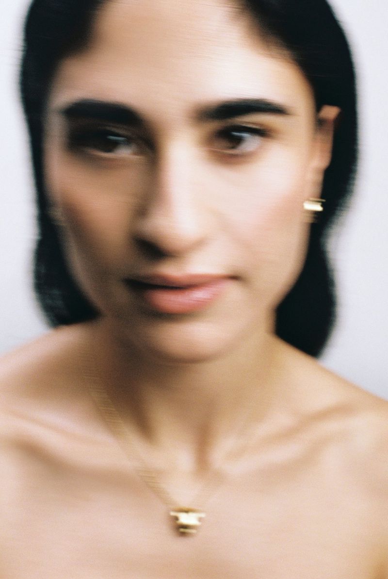 Close-up portrait of a woman with dark hair and minimal jewelry, featuring a gold necklace and earrings against a soft background.