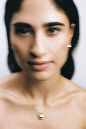 Close-up portrait of a woman with dark hair wearing a gold pendant necklace and earrings against a pale background.