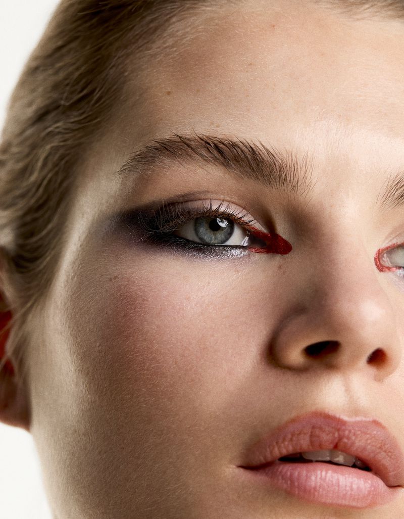 Close-up of a woman's face with dramatic makeup, featuring dark eyeshadow and red accents, against a neutral white background.