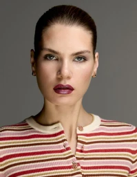 Close-up beauty portrait of a woman with slicked-back hair and metallic berry lipstick wearing a striped knit cardigan.