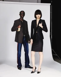 Two fashion models standing in studio wearing tailored blazers, dark sunglasses, denim jeans, and a black skirt suit.