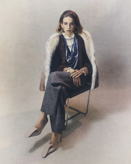 A young woman in a fur-collared coat sits on a metal chair wearing grey tweed pants and light-beige heels against a neutral backdrop.