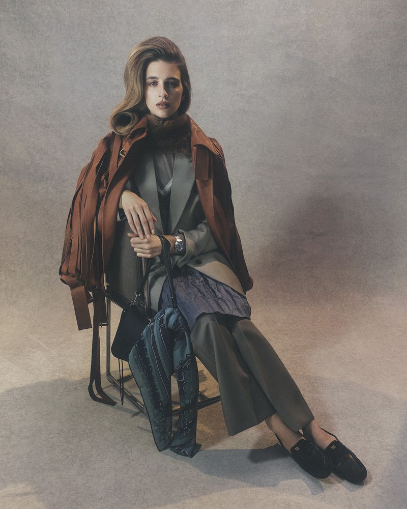 Elegant woman poses seated in tailored coat and pants, accessorized with a scarf, jacket, handbag, and loafers against a grey backdrop.