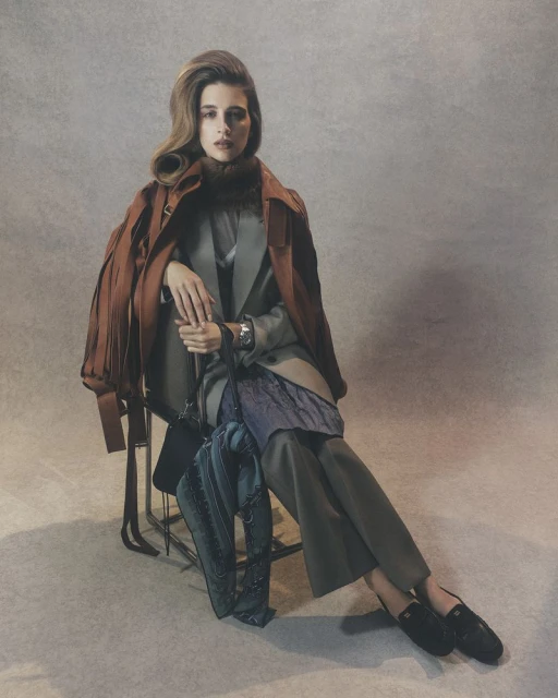 Elegant woman poses seated in tailored coat and pants, accessorized with a scarf, jacket, handbag, and loafers against a grey backdrop.
