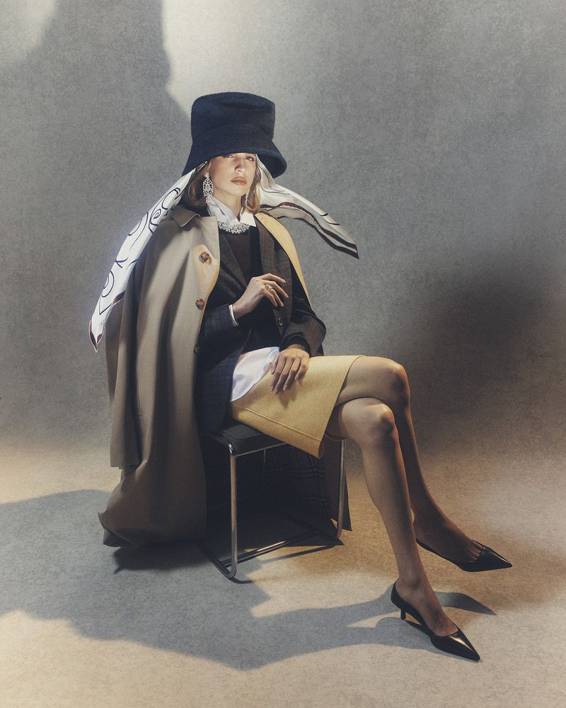 Stylish woman in a black hat and beige trench coat sits on a chair in a studio with soft lighting, showcasing fashion and elegance.