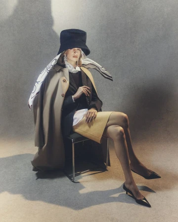 Stylish woman in a black hat and beige trench coat sits on a chair in a studio with soft lighting, showcasing fashion and elegance.