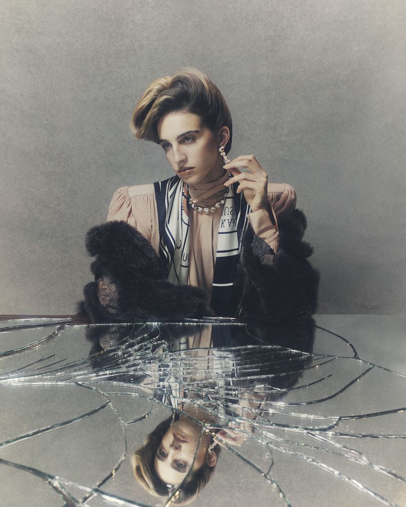 Woman with a strong makeup look wearing statement earrings, a pearl necklace, and a fur stole reflected in a broken mirror.