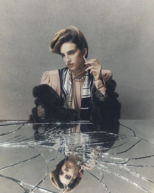 Woman with a strong makeup look wearing statement earrings, a pearl necklace, and a fur stole reflected in a broken mirror.