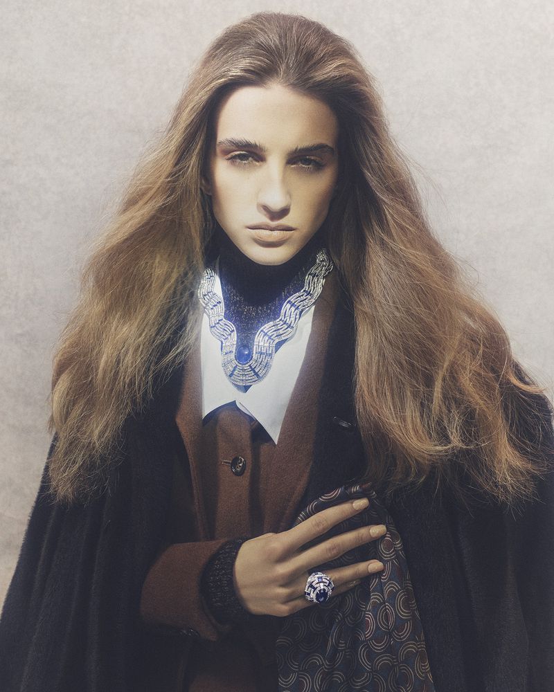 Fashion photo of a woman with long brown hair wearing a blue and silver necklace, brown jacket, and black cape, posing indoors.