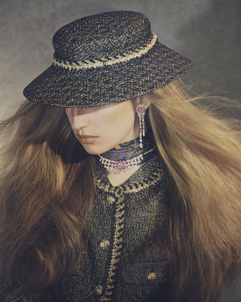 Fashion portrait of a woman wearing a wide-brimmed hat, jacket, and jewelry with long blonde hair against a muted grey studio backdrop.