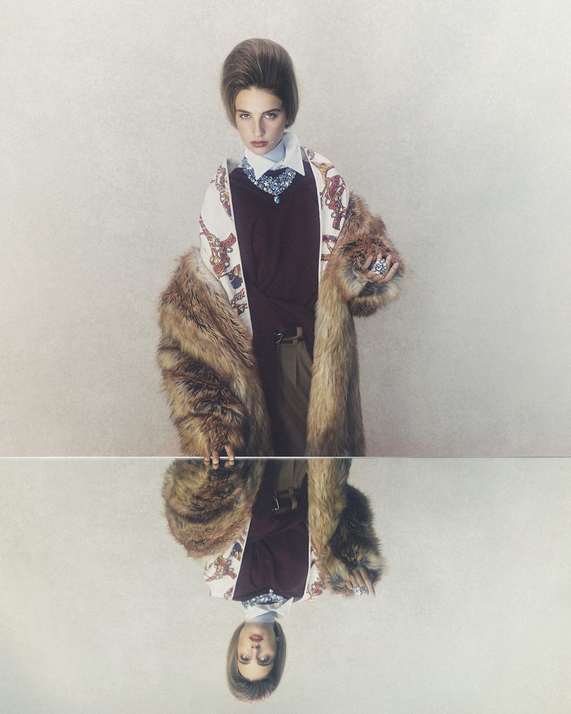 Fashionable woman poses in a studio, wearing a fur coat, sweater, patterned blouse, and skirt, reflected in a mirror surface below.