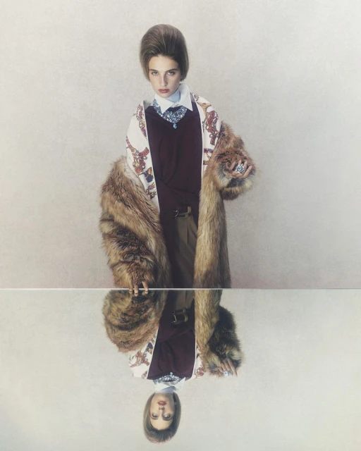 Fashionable woman poses in a studio, wearing a fur coat, sweater, patterned blouse, and skirt, reflected in a mirror surface below.