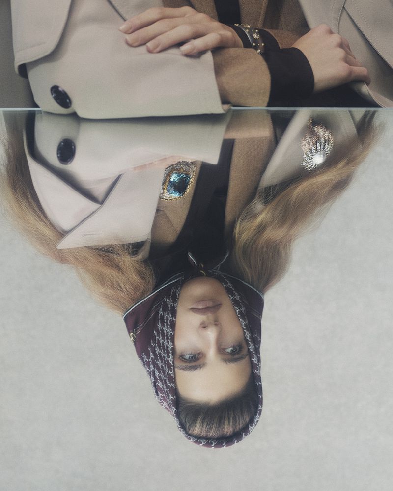 Woman wearing a patterned headscarf and a tan trench coat is reflected in a glass surface, offering a mirrored perspective.