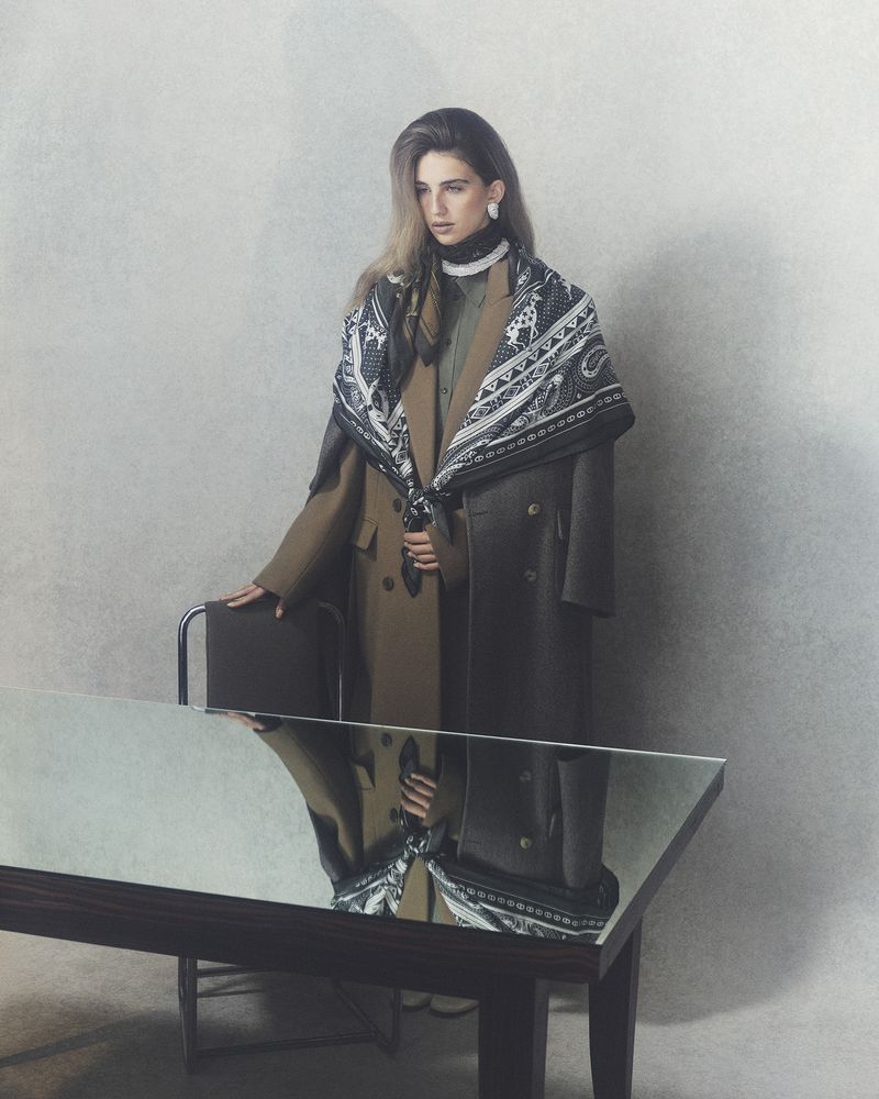 Fashionable adult female in double-breasted coats and paisley scarf poses with chair, reflected in a sleek black table in a photo studio.