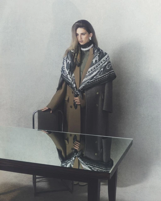 Fashionable adult female in double-breasted coats and paisley scarf poses with chair, reflected in a sleek black table in a photo studio.