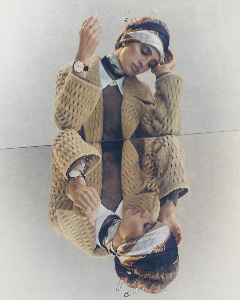 Fashion shot of a woman wearing a beige patterned jacket, white shirt, brown sweater and headscarf, reflected in a mirror.
