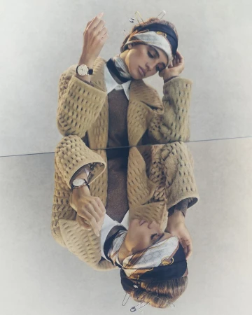 Fashion shot of a woman wearing a beige patterned jacket, white shirt, brown sweater and headscarf, reflected in a mirror.
