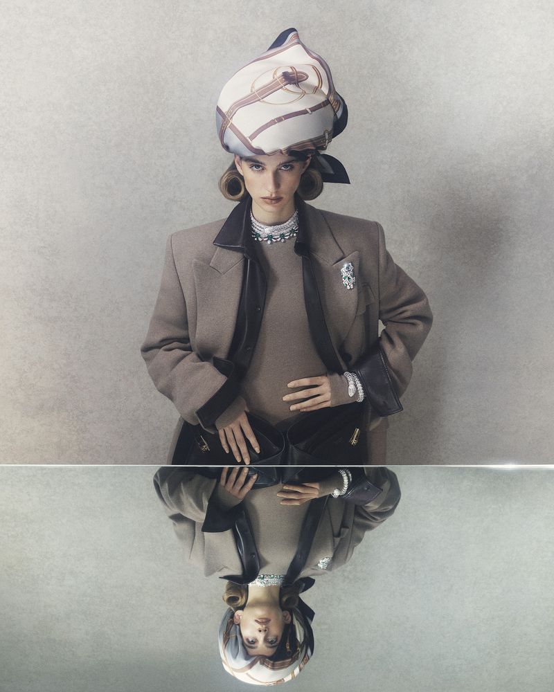 Model Lota Blaskovic wearing a tailored beige coat and patterned headscarf, reflected in a horizontal mirror surface.