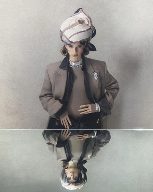 Model Lota Blaskovic wearing a tailored beige coat and patterned headscarf, reflected in a horizontal mirror surface.