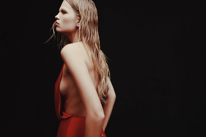 Blonde woman in a red dress stands against a dark backdrop, showcasing a side profile with wet hair and a minimalist aesthetic.