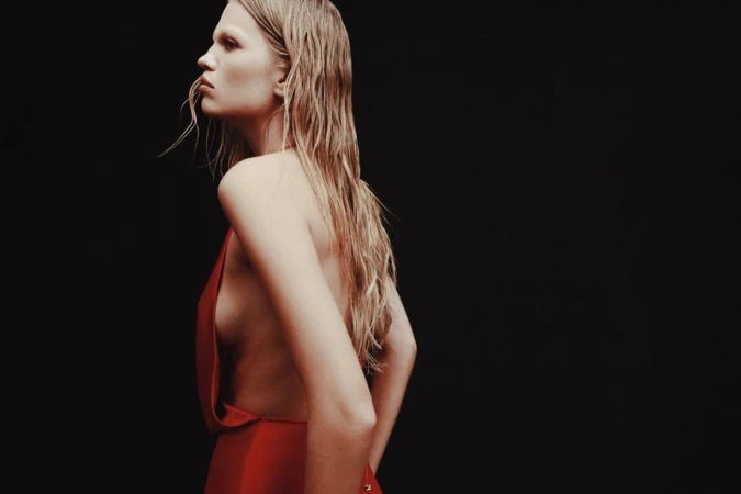 Blonde woman in a red dress stands against a dark backdrop, showcasing a side profile with wet hair and a minimalist aesthetic.