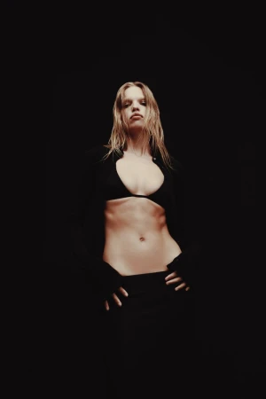 A fair-skinned adult female model with wet blonde hair poses in a black bikini top and pants against a dark background, gazing confidently.