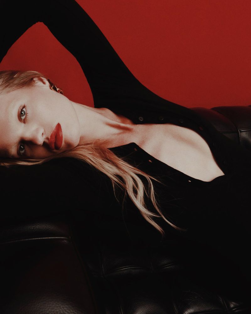 Blonde woman in dark clothing reclines on a black leather sofa against a red background, with red lipstick.