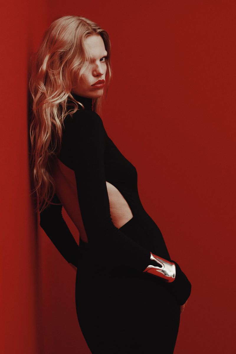 A fair-skinned woman with blond, wavy hair poses against a red wall, wearing a black dress with an open back and silver cuff.