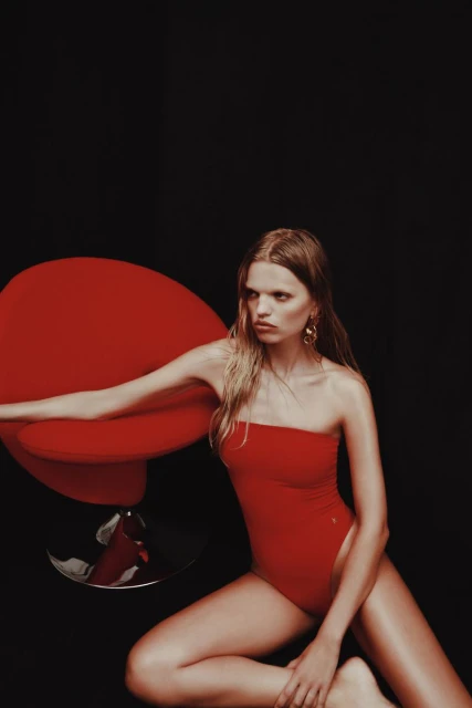 Woman with long blonde hair poses in a red swimsuit next to a red chair against a black background. Fashion editorial shot.
