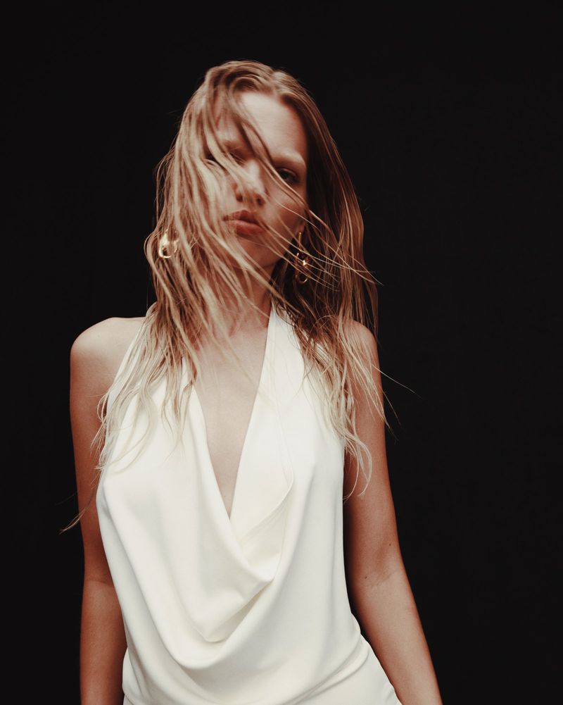 Close-up portrait of a woman with wet blonde hair and a white halter top against a dark background, showcasing fashion and beauty.