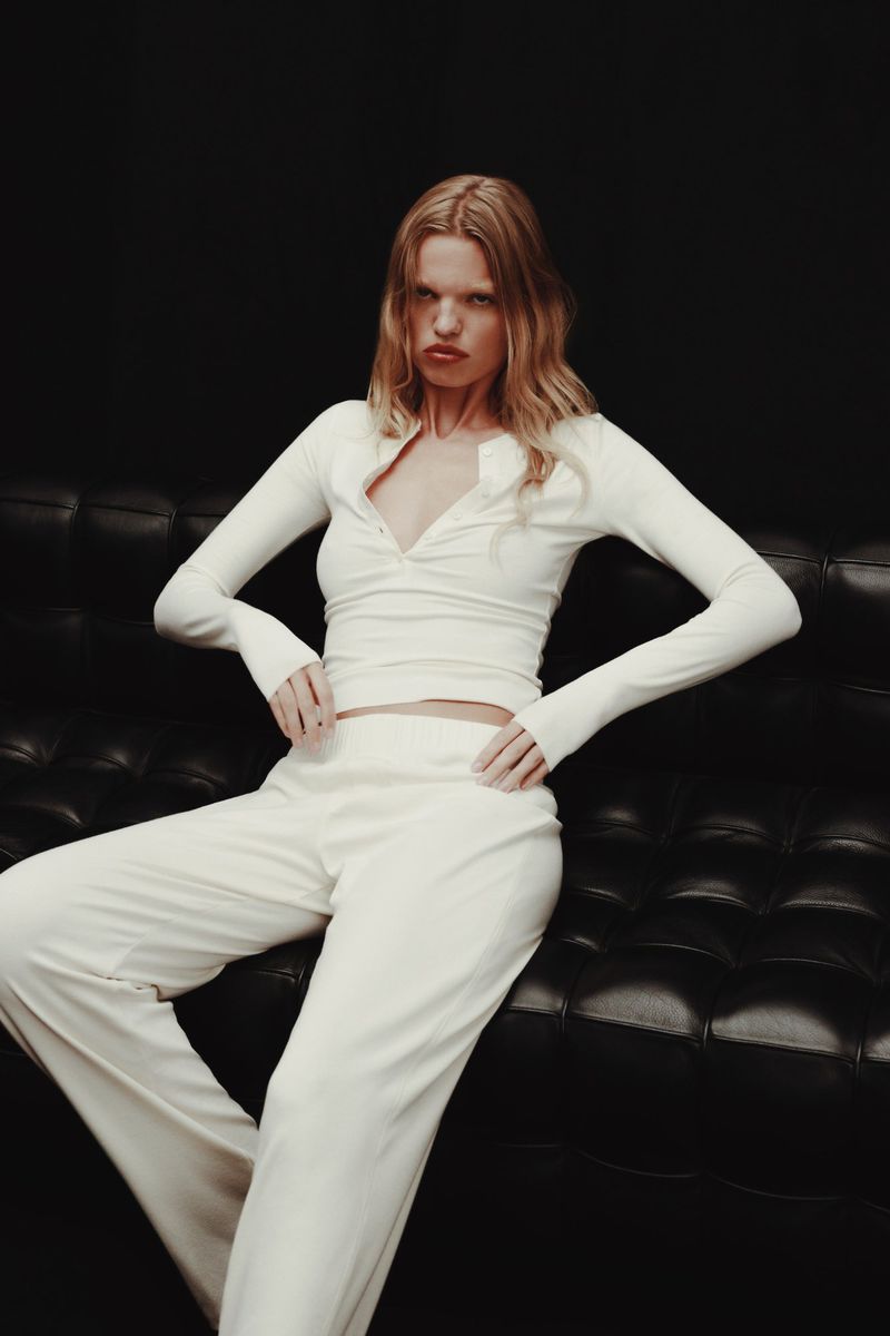 Fashion portrait of a blonde woman in a white long-sleeved top and white pants, sitting on a black leather couch against a dark background.