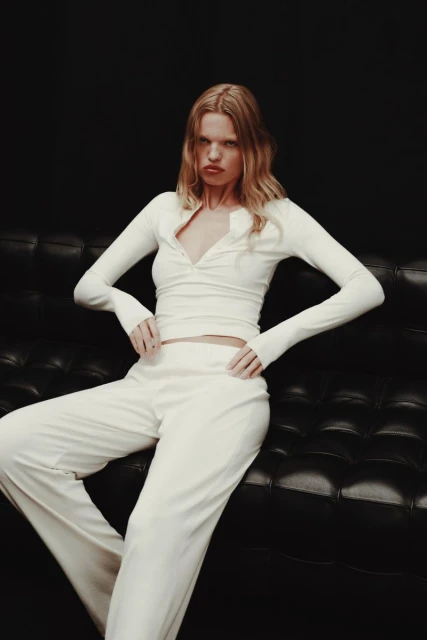 Fashion portrait of a blonde woman in a white long-sleeved top and white pants, sitting on a black leather couch against a dark background.
