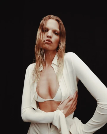 Model Daphne Groeneveld wearing an unbuttoned cream shirt and matching bikini top against a solid black backdrop.