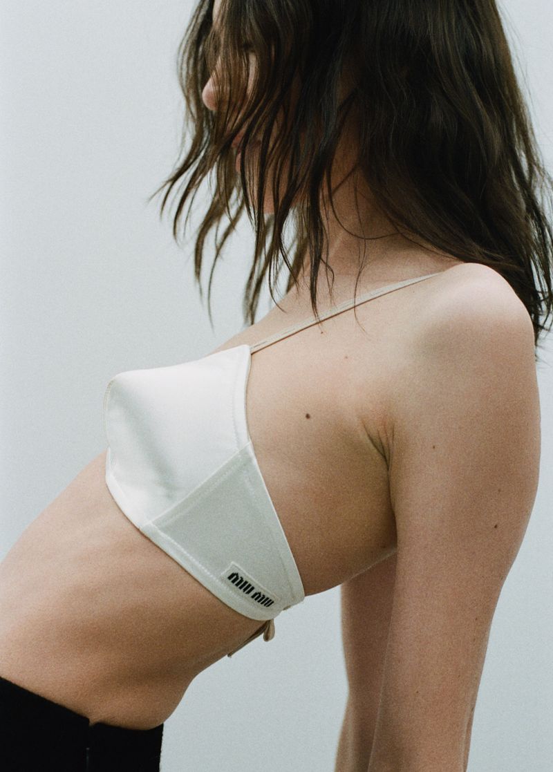 Brunette adult female model poses in a white designer bra top with a visible Miu Miu logo against a neutral studio background.
