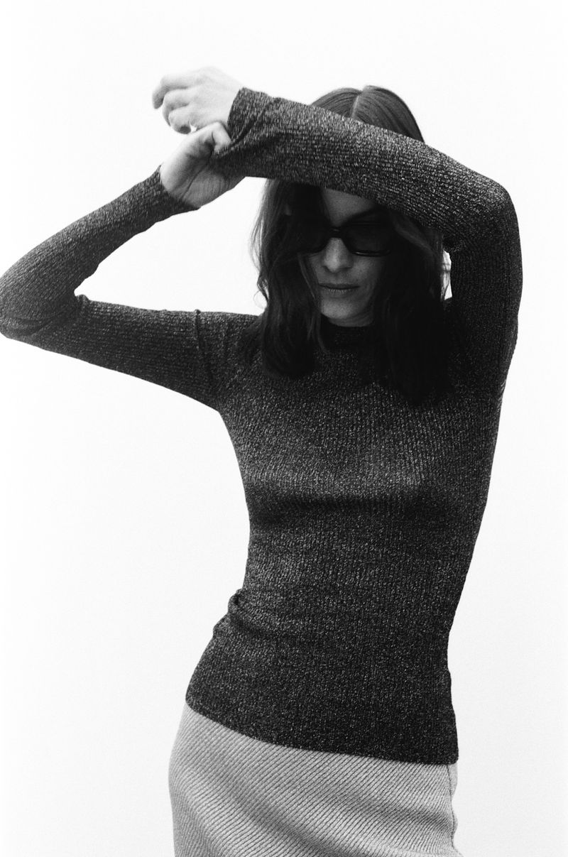 A woman poses indoors with her arms above her head, wearing sunglasses and a dark shimmering sweater with a grey skirt, against a white backdrop.
