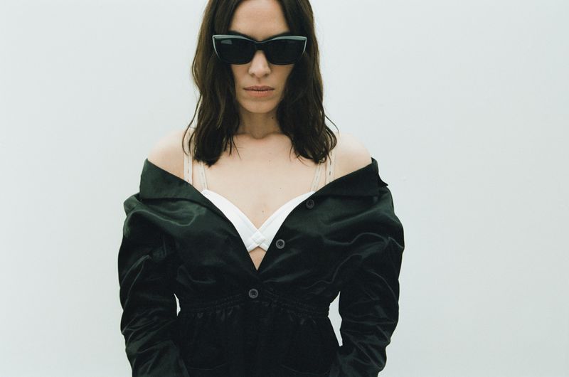 Woman with dark hair and sunglasses poses in a white top and unbuttoned black jacket against a clean white background in a studio shot.