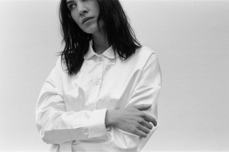 Monochrome portrait of a stylish adult woman with dark hair, wearing a white collared shirt, with arms crossed, looking left, in a photo studio.