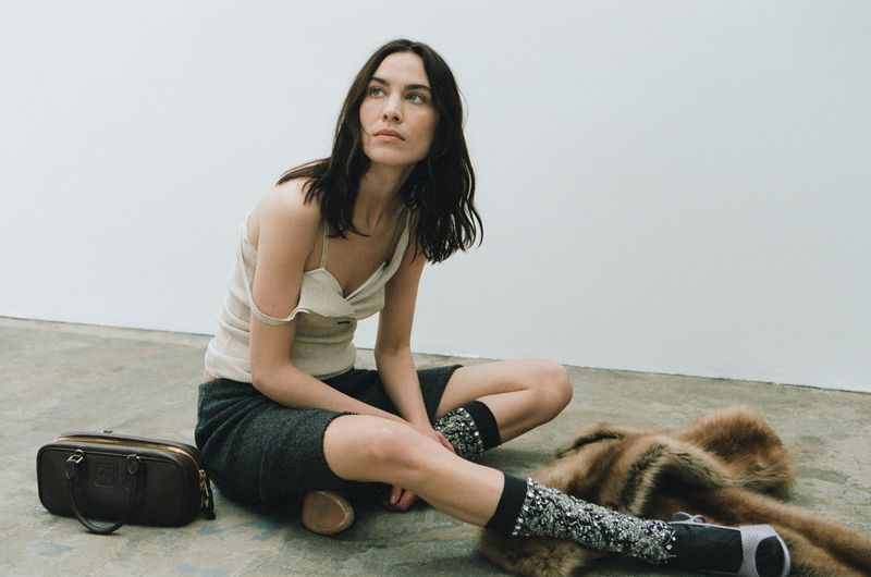 A woman sits indoors in a cream tank top and dark shorts, with a brown bag, patterned socks, and a fur stole visible.