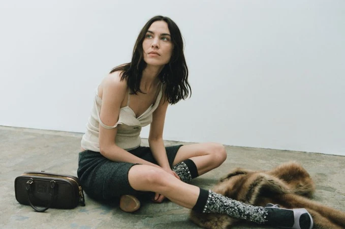 A woman sits indoors in a cream tank top and dark shorts, with a brown bag, patterned socks, and a fur stole visible.