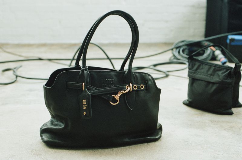 Black leather tote bag with gold hardware and a tag, positioned on a light grey surface with black electrical cables in the background.