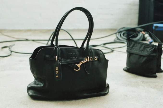 Black leather tote bag with gold hardware and a tag, positioned on a light grey surface with black electrical cables in the background.