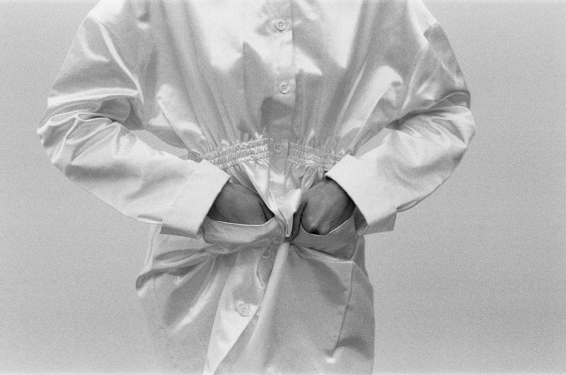 Person in white shirt with hands in pockets, black and white fashion shot. Simple background, monochrome aesthetic, relaxed posing, bright lighting.