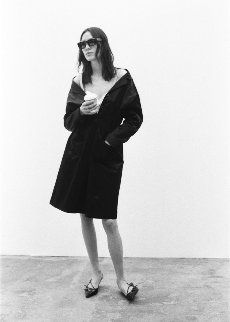 Monochrome studio shot of an adult female in a coat and sunglasses holding a beverage cup, standing on a gray concrete floor.