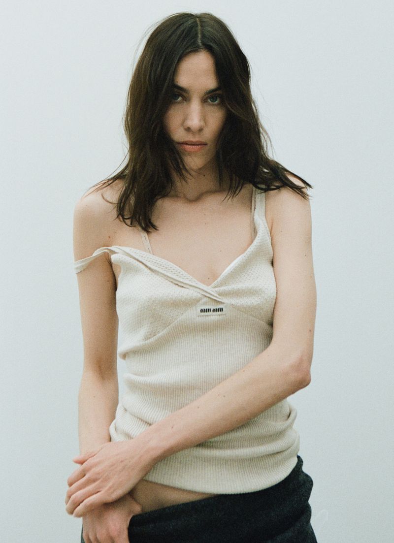 Brunette woman in a cream ribbed tank top with thin straps and a black skirt or pants, posing in front of a pale neutral background.