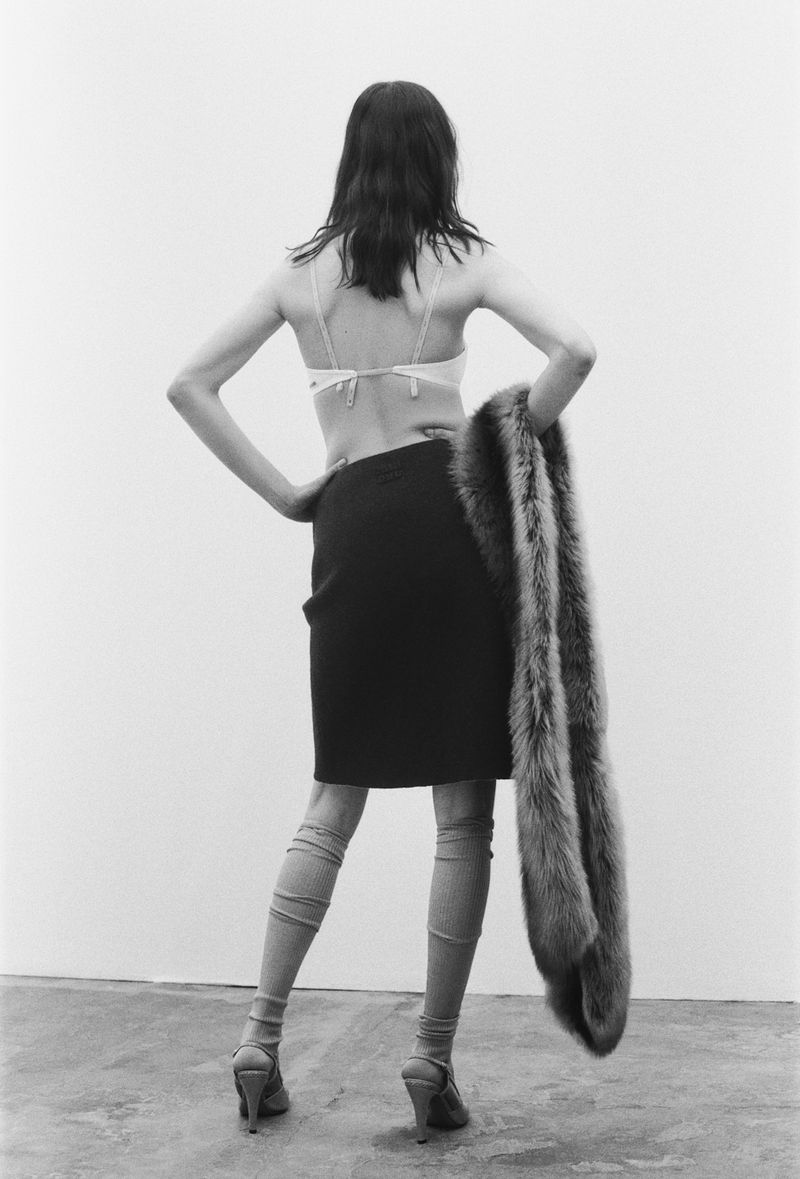 Back view of a slim adult woman in a bra top and skirt, standing in a minimalist photo studio, holding a fur stole over her shoulder.