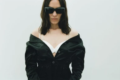 Alexa Chung wearing dark cat-eye sunglasses and an off-the-shoulder black jacket over a white top against a minimal pale backdrop.