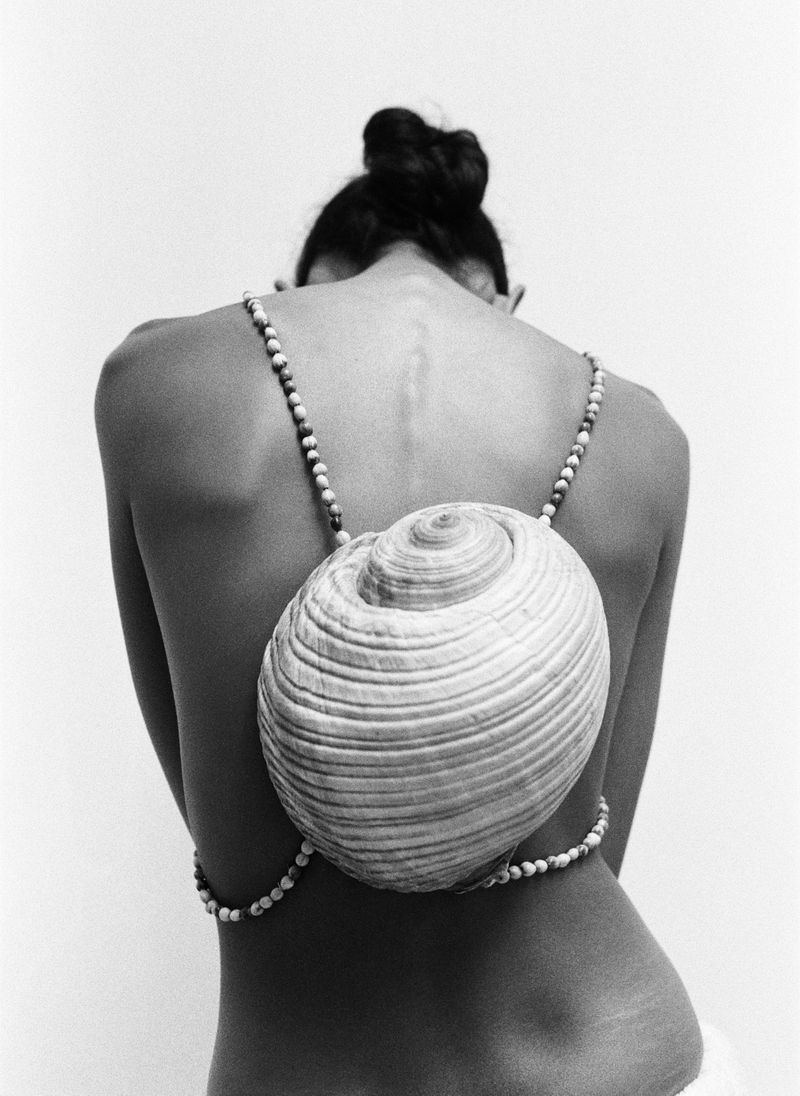 Back view of woman wearing large seashell and beaded strings, set against a stark white backdrop in a black and white photograph.