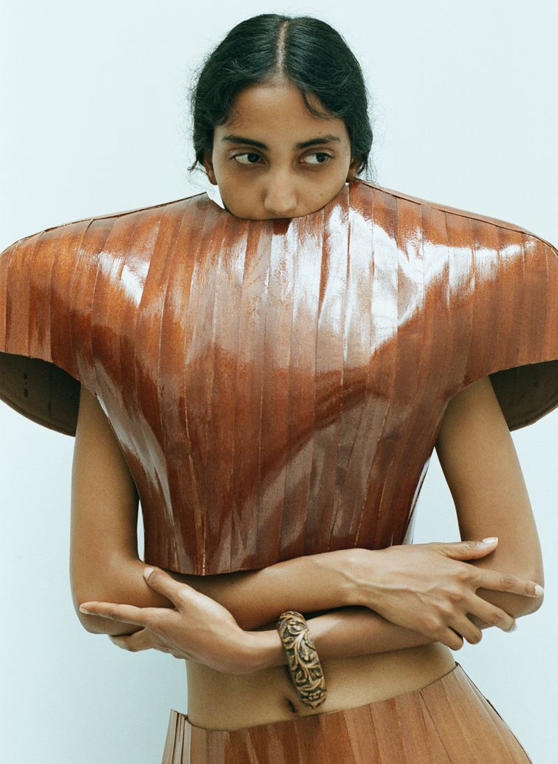 Adult woman in a sculptural wood top and skirt poses in a studio with her arms crossed, creating a unique editorial fashion portrait.