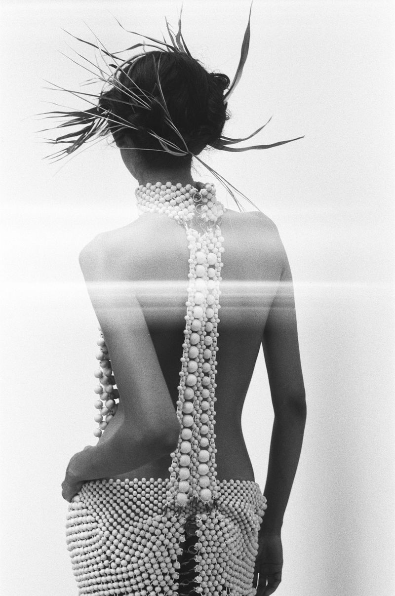 A woman with an artistic feathered headpiece stands with her back to the camera wearing beaded dress and necklace against a white backdrop.
