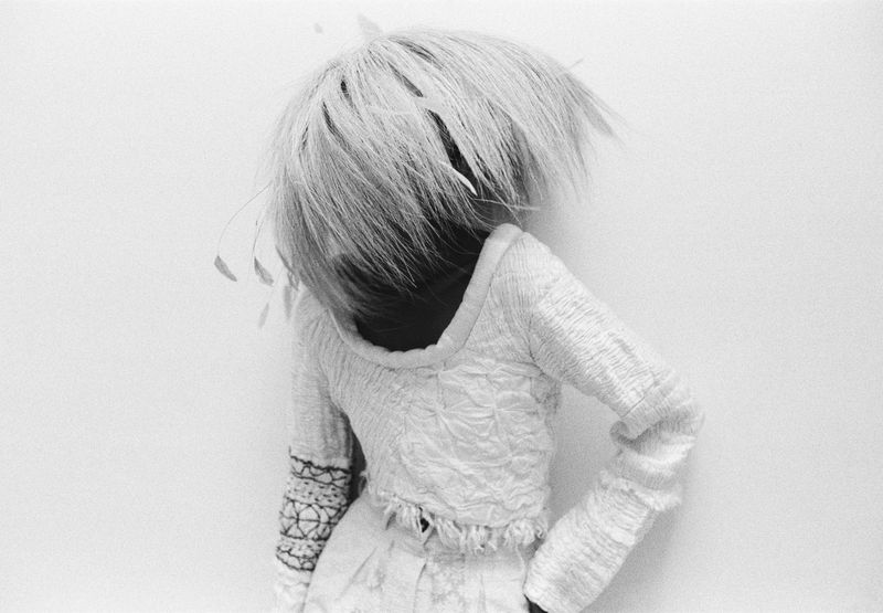 Person in textured white outfit with voluminous light hair against a plain white wall, black and white fashion photography.